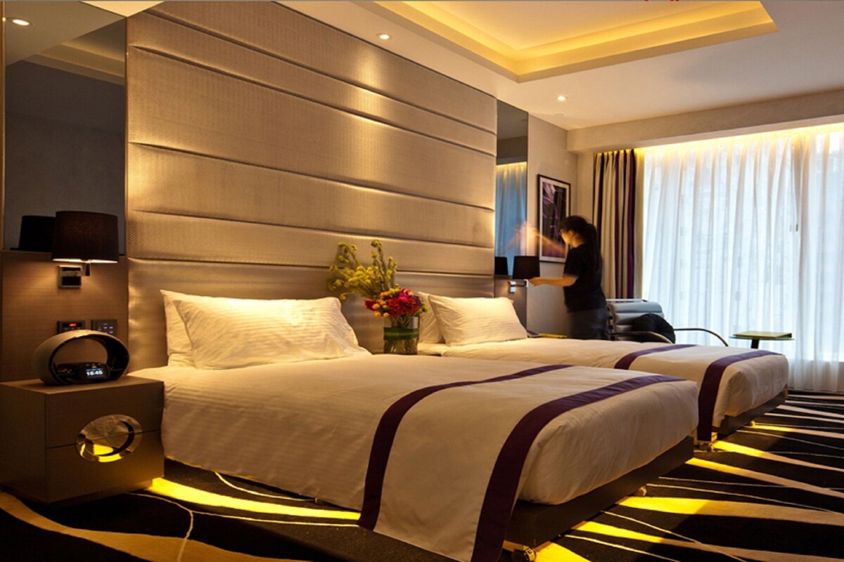 A hotel room with two beds, a nightstand with a clock, a small table, a chair, and a large window with curtains.