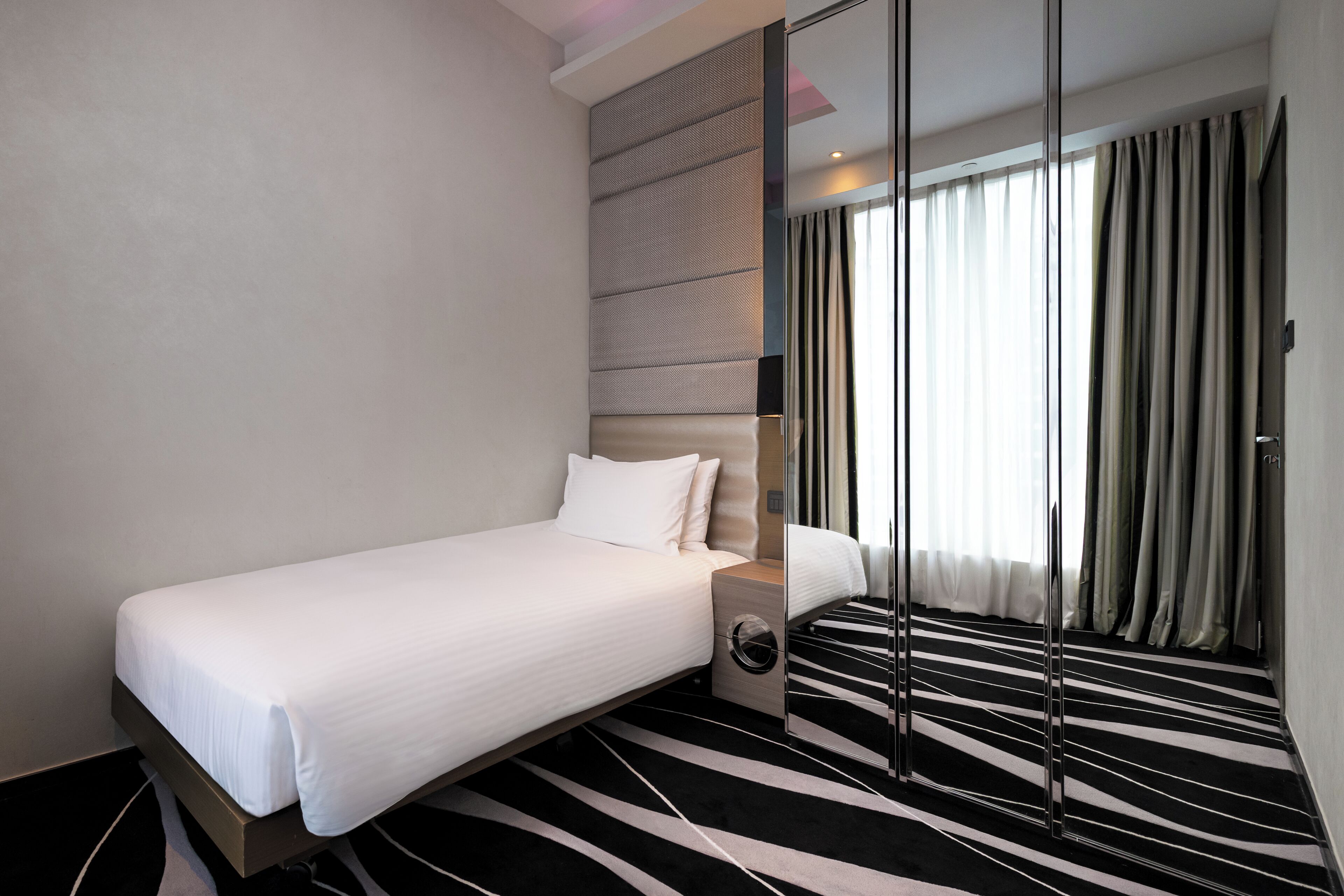 A hotel room with a bed, a bedside table, a mirror, a window with curtains, and a striped carpet.