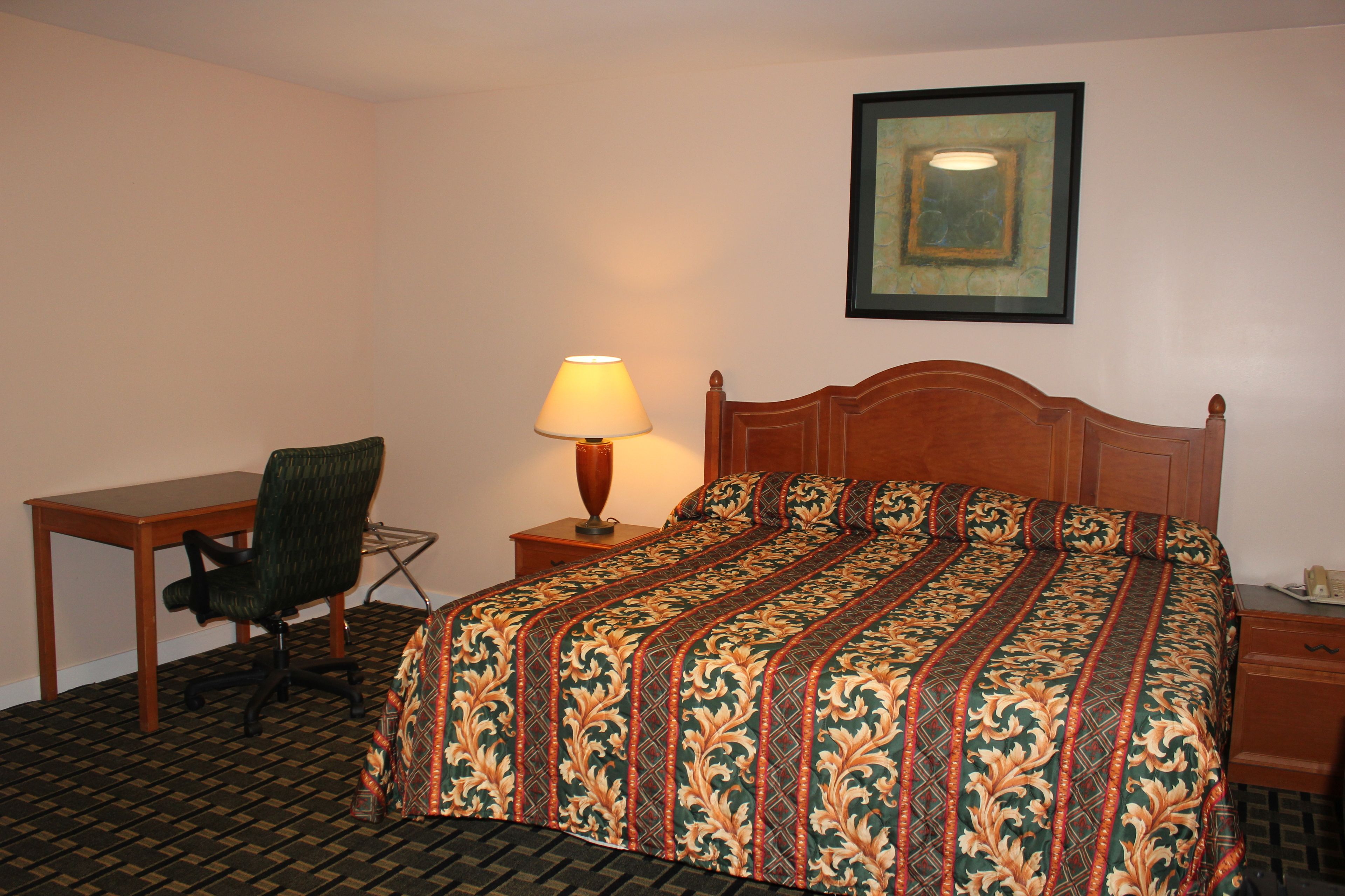A hotel room with a wooden bed, a desk, a chair, a lamp, and a framed picture on the wall.