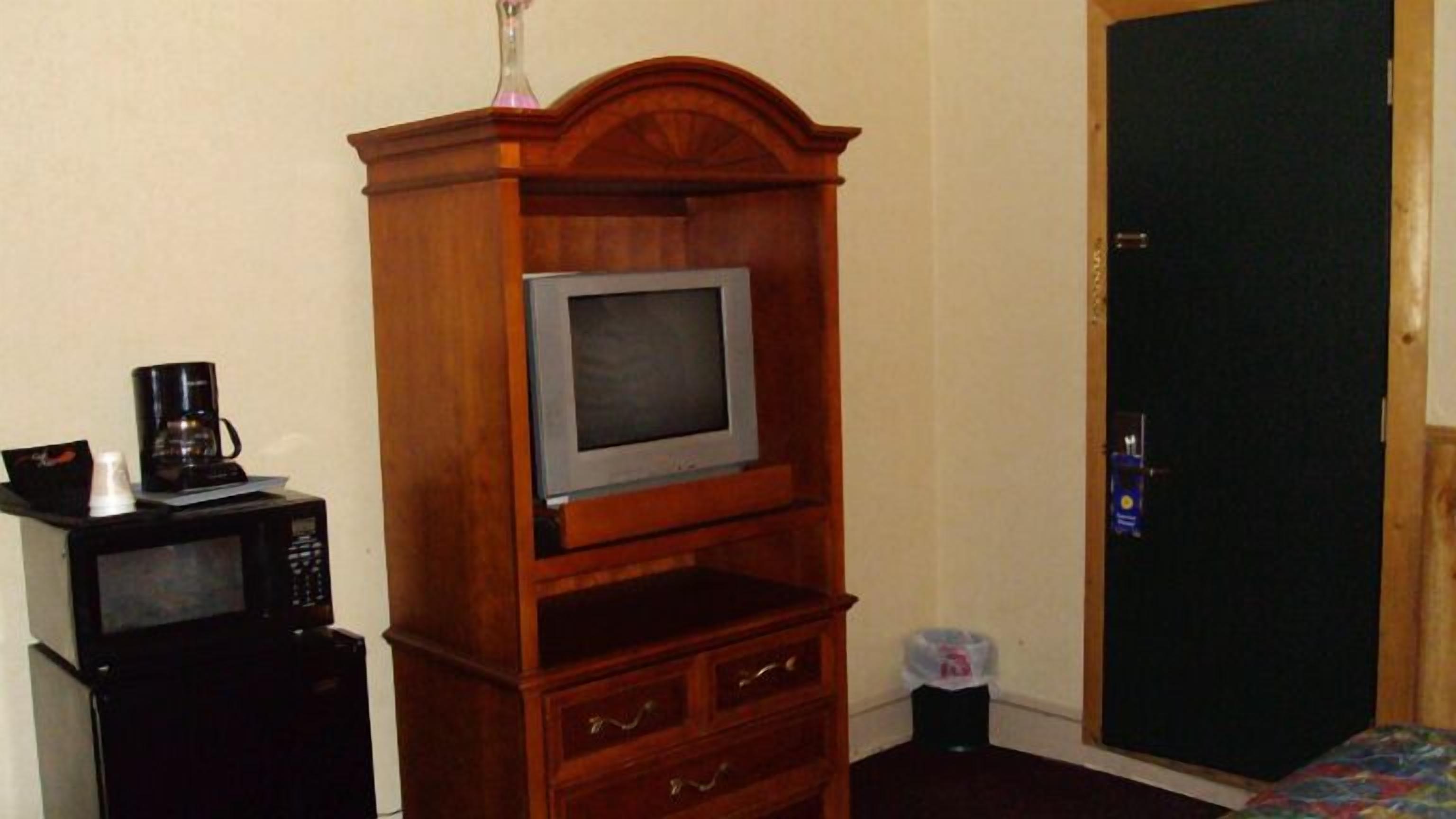Una stanza con un mobile in legno contenente un televisore, una macchina del caffè e un forno a microonde. Si apre una porta a destra ed è presente un cestino nell'angolo.