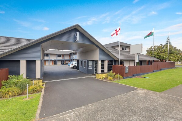 A modern building with a covered entrance, a parking area, and flags. - ASURE Aveda Motor Lodge (Pukekohe)