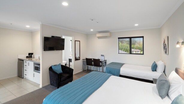 A hotel room with two beds, a desk, a chair, a TV, and a kitchenette. - ASURE Aveda Motor Lodge (Pukekohe)