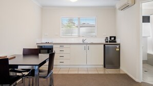A compact kitchen with a dining table, chairs, a refrigerator, and a window with blinds. - ASURE Aveda Motor Lodge (Pukekohe)