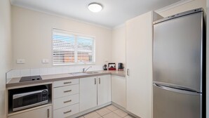 A modern kitchen with white cabinets, a stainless steel refrigerator, a microwave, and a window with blinds. - ASURE Aveda Motor Lodge (Pukekohe)