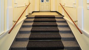 A grand staircase with a carpeted finish, flanked by white walls and columns, leading to a door with a glass panel. - Alqush Downtown Hotel (Prague)