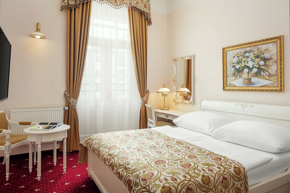 A hotel room with a bed, a desk with a chair, a small table, a lamp, and a painting on the wall.