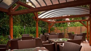 An outdoor patio with wicker furniture and glass tables, surrounded by a wooden pergola and greenery. - Alqush Downtown Hotel (Prague)
