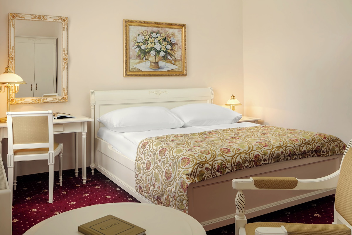 A hotel room with a bed, a desk, a chair, a round table, a mirror, and a painting on the wall.