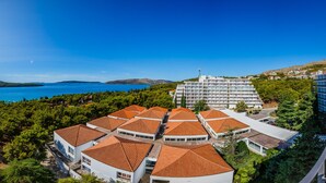 A coastal hotel with multiple buildings, a prominent white structure with a red roof, and a view of the sea and hills in the background. -  Hotel Medena Budget (Seget)