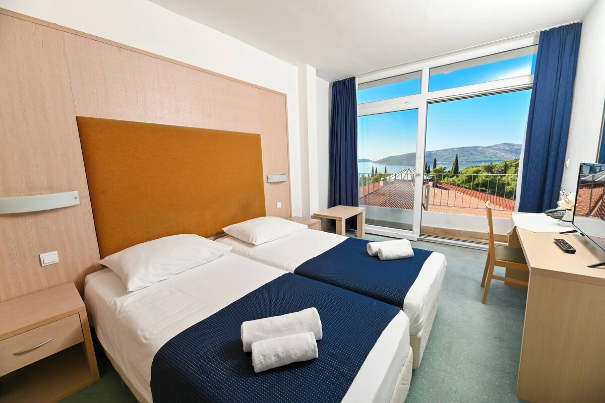 Habitación de hotel con dos camas, un escritorio, una silla y un ventanal con vistas a las montañas.