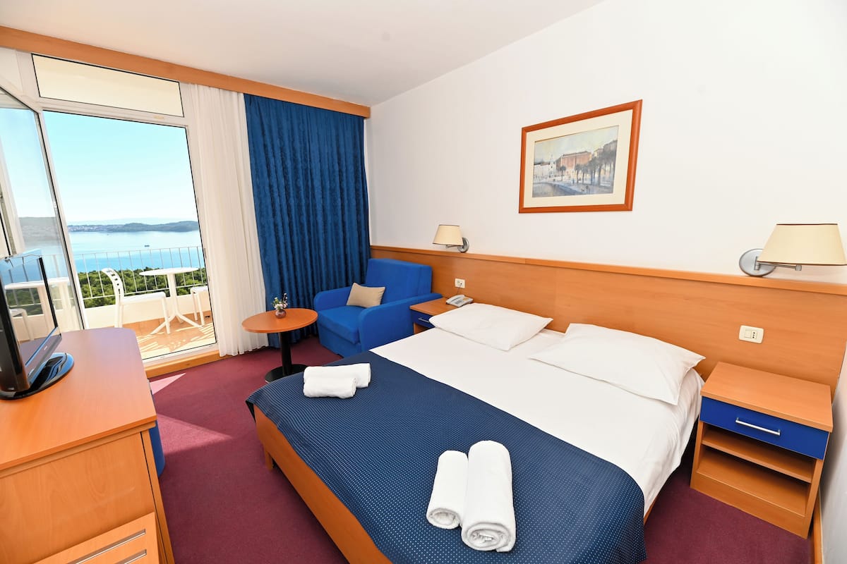 Habitación de hotel con cama, escritorio, silla, mesita, mesita de noche y ventana con vista al mar.