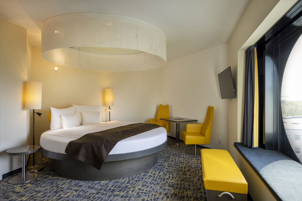 Una habitación de hotel moderna con una cama curvada, una mesa redonda, una silla amarilla, un banco y un televisor montado en la pared.