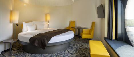 A modern hotel room with a curved bed, a round table, a yellow chair, a bench, and a TV mounted on the wall.