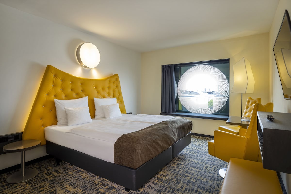 Habitación de hotel con una cama grande, una lámpara redonda fijada a la pared, una ventana con vistas, un escritorio y una silla.