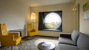 A hotel room with a sofa, two yellow armchairs, a small table, a lamp, and a large window with a view of greenery.