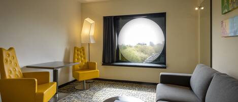 Habitación de hotel con sofá, dos sillones amarillos, una mesita, una lámpara y un amplio ventanal con vistas a zonas verdes.