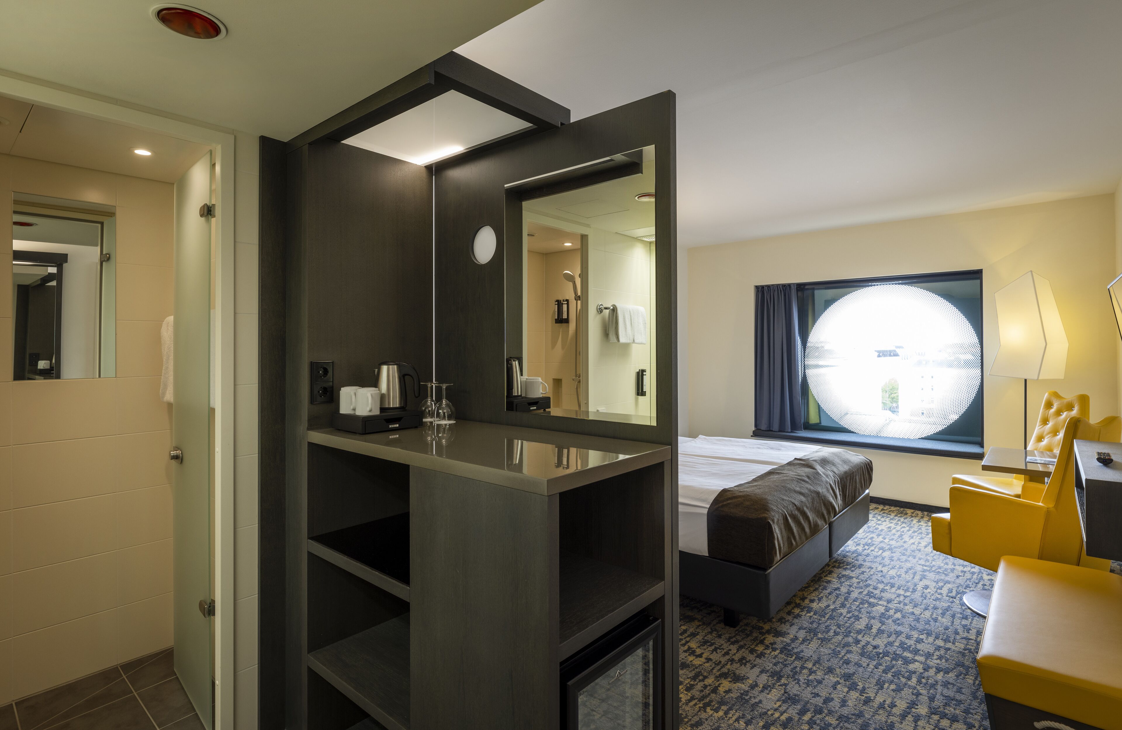 A modern hotel room with a large bed, a bedside table, a mirror, a window with a view, and a small seating area.