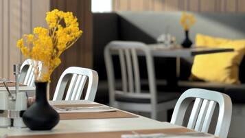 A dining area with white chairs, a table with a vase of yellow flowers, and a sofa with yellow cushions.