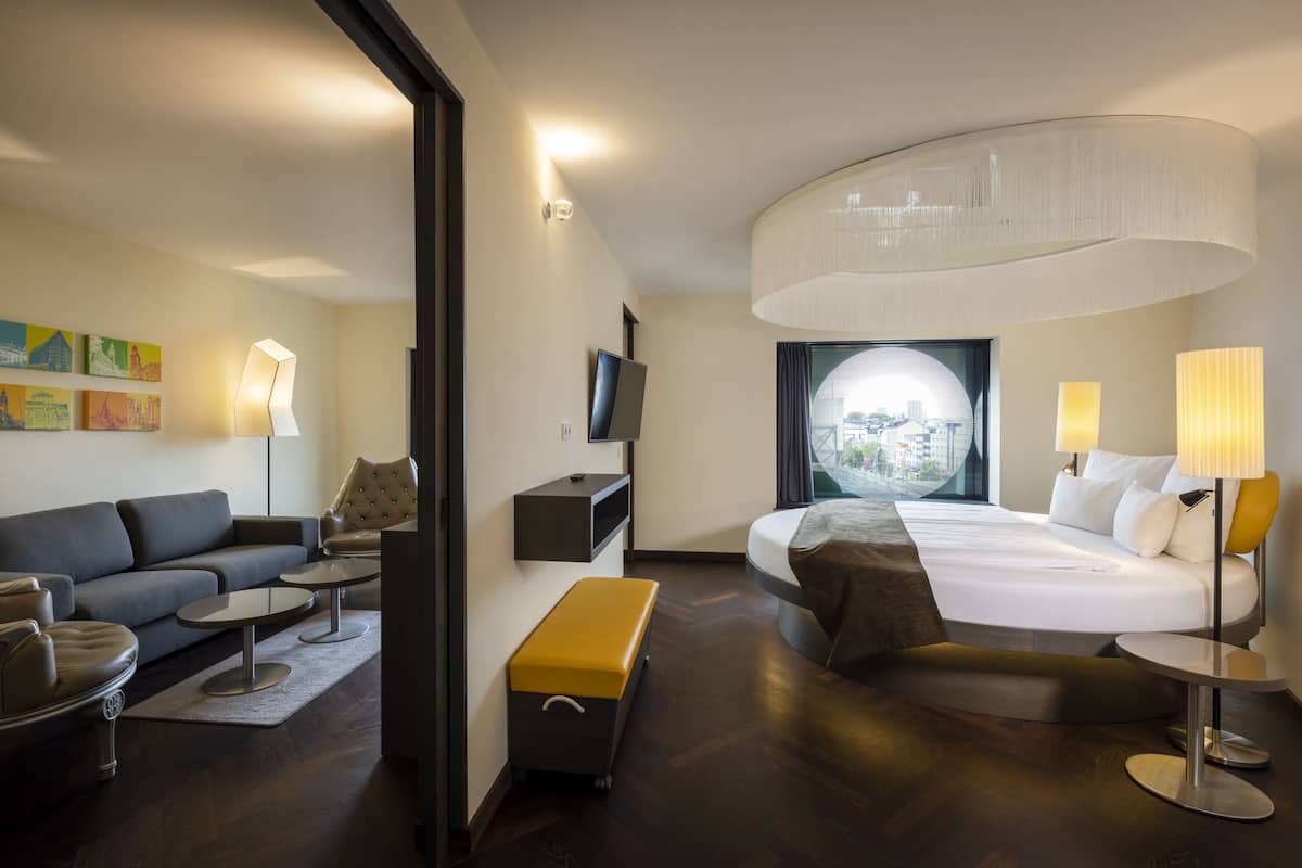 Una habitación de hotel moderna con techo curvo, una cama grande, un sofá, una mesa de centro, una banca y un televisor.
