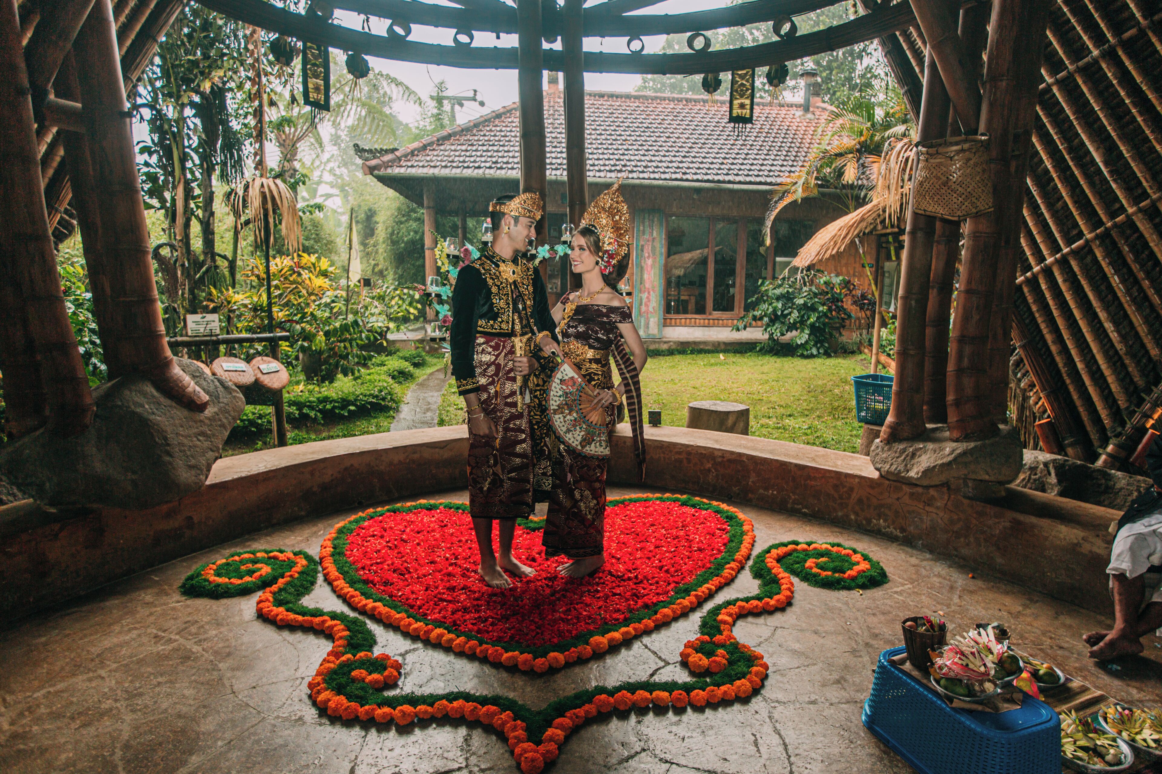 Un couple se tient dans un espace décoré, avec un motif en forme de cœur sur le sol, entouré de verdure et d’un bâtiment traditionnel.