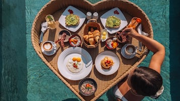 A heart-shaped tray with various breakfast items and drinks, placed on a poolside table.