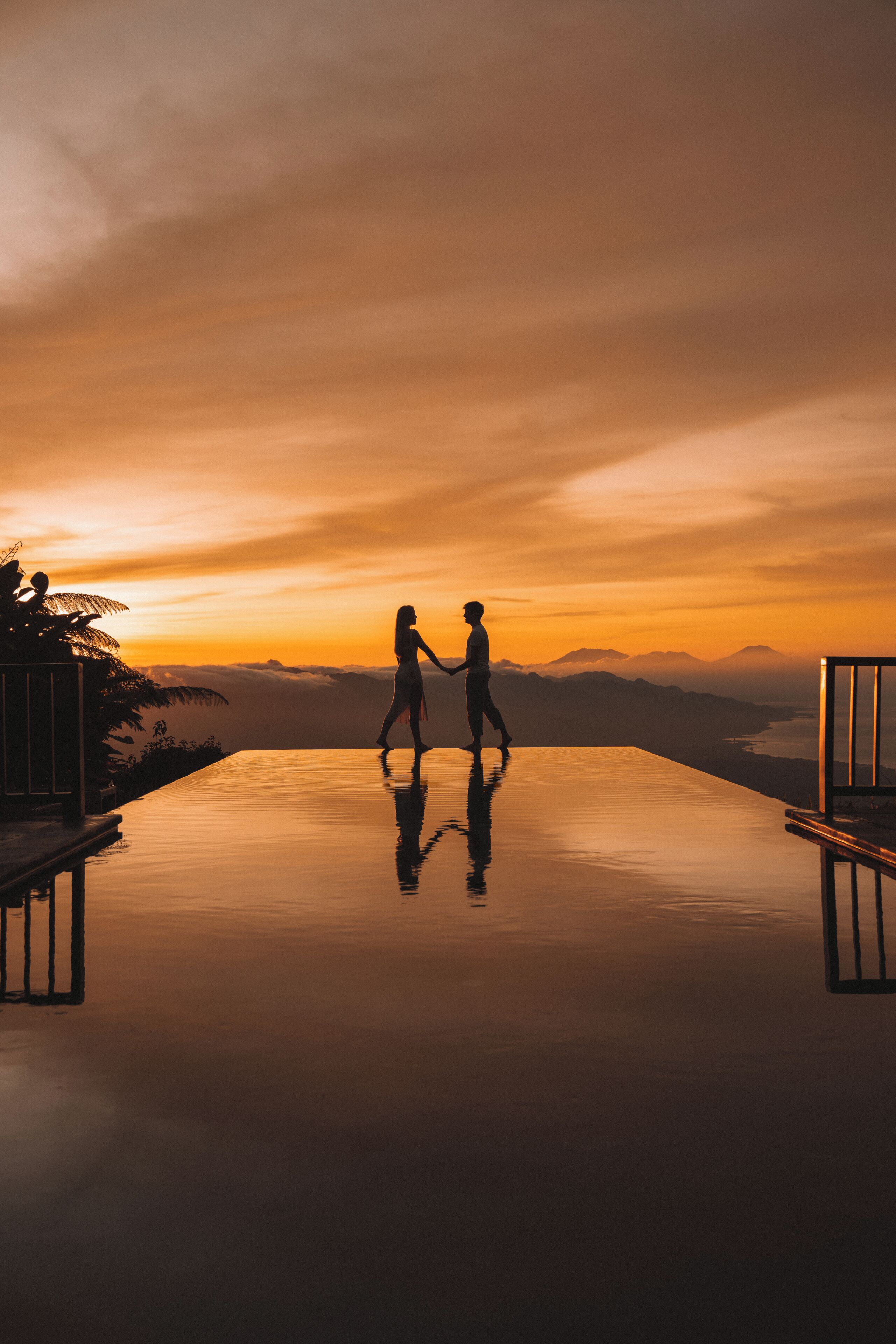 A couple walking on a reflective infinity pool with a sunset view.