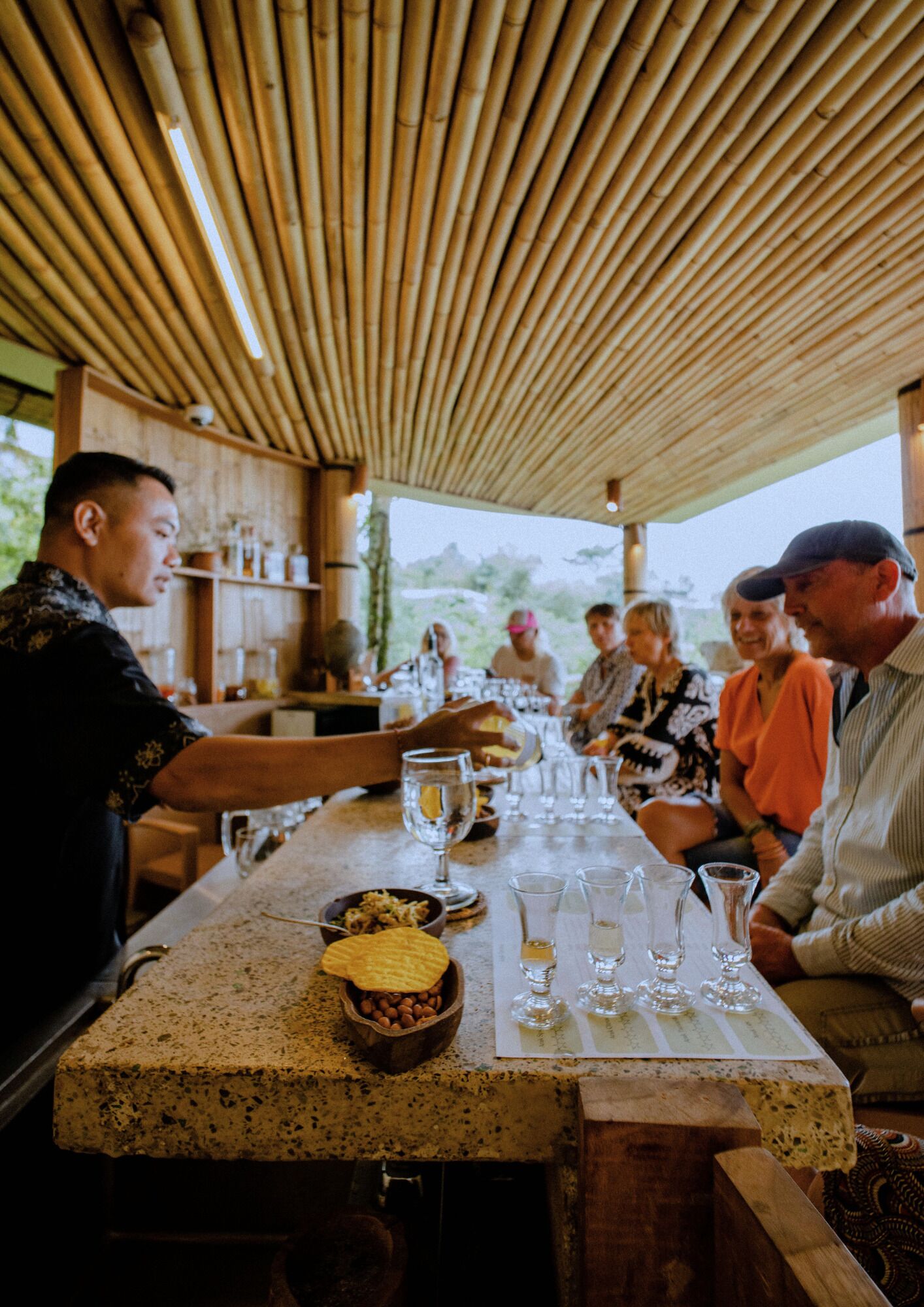 Un groupe de personnes est assis à une table, sous une pergola recouverte, avec un plafond en bambou, et peut admirer la verdure environnante.