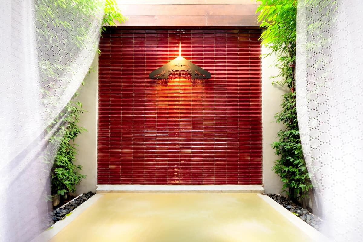 Une porte rouge ornée d’un luminaire décoratif suspendu, entourée de verdure et agrémentée d’une petite fontaine.