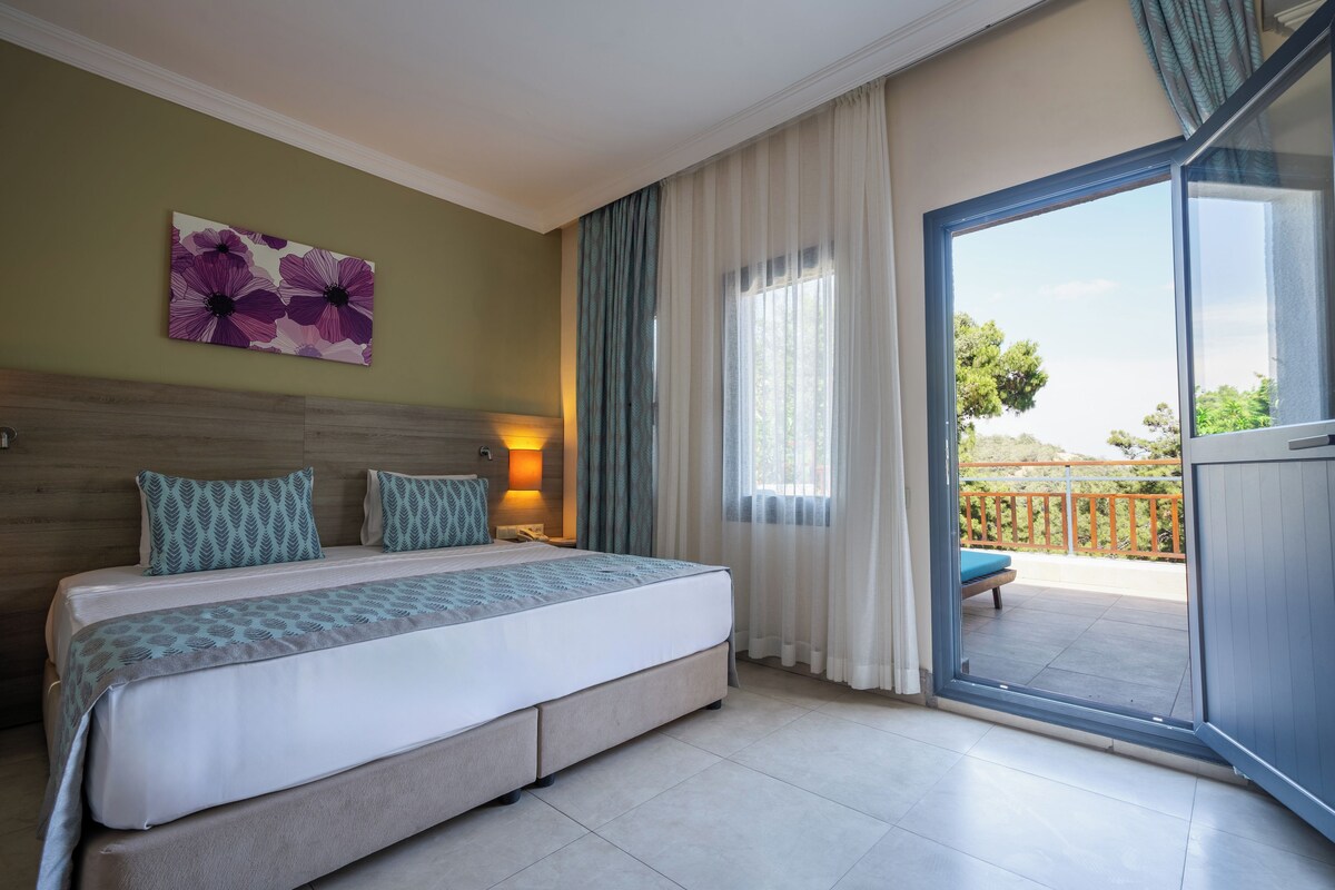 Habitación de hotel con cama, mesita de noche, lámpara, ventana con cortinas y vistas al exterior.
