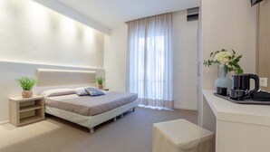 Una habitación de hotel moderna con una cama grande, una mesita de noche con una planta, un pequeño rincón de descanso y una kitchenette con cafetera.