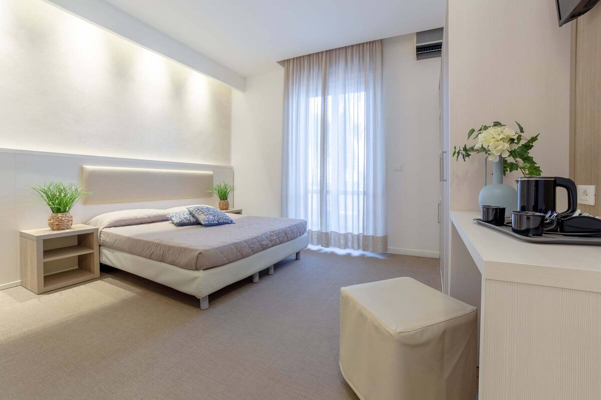 Une chambre d’hôtel moderne dotée d’un grand lit, d’une table de chevet avec une plante, d’un petit coin salon et d’une kitchenette équipée d’une machine à café.