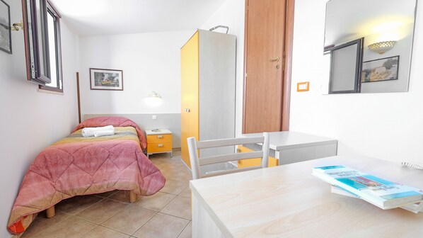 Un dormitorio con una cama, un escritorio y una silla. Incluye un armario, una mesita de noche y una lámpara.