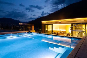 Une piscine avec une paroi en verre, un espace détente avec des sièges et une vue sur les montagnes en arrière-plan.