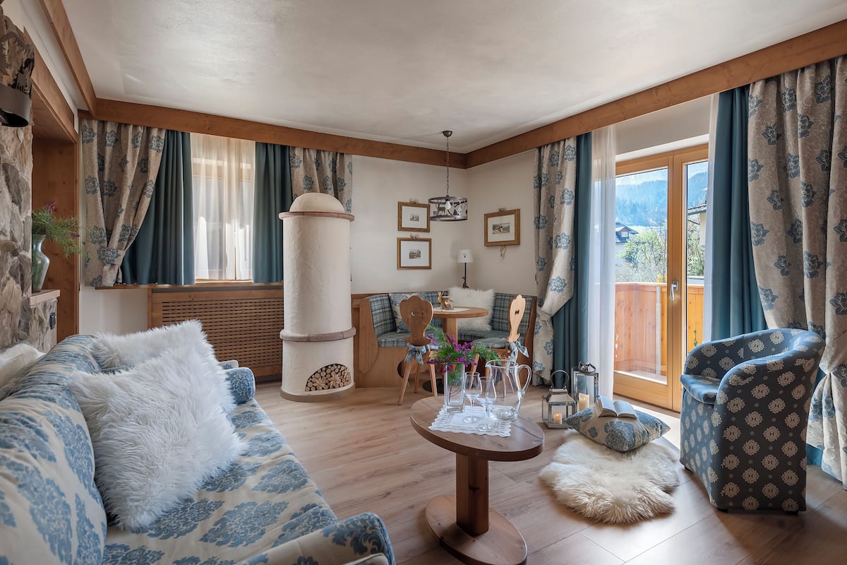 Ein gemütliches Wohnzimmer mit einer Couch, einem Sessel und einem runden Tisch mit Gläsern. Außerdem ist eine Holzbank und ein kleiner Tisch mit Blumenvase zu sehen.