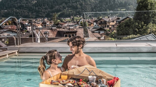 Un couple se trouve dans une piscine, près d’une table en bois où sont disposés des plats et des boissons. Ils sont entourés d’un paysage montagneux, et on aperçoit une ville au loin.