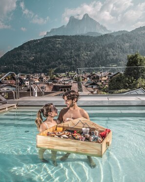 Un couple se trouve dans une piscine, près d’une table en bois où sont disposés des plats et des boissons. Ils sont entourés d’un paysage montagneux, et on aperçoit une ville au loin.