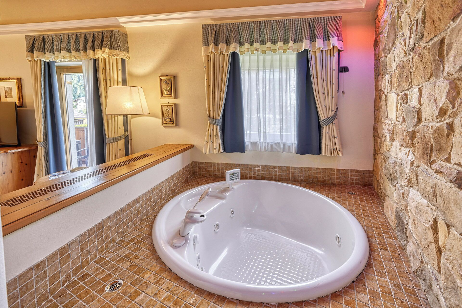 Une salle de bain avec une baignoire, une fenêtre avec des rideaux, un banc en bois et un mur en pierre.