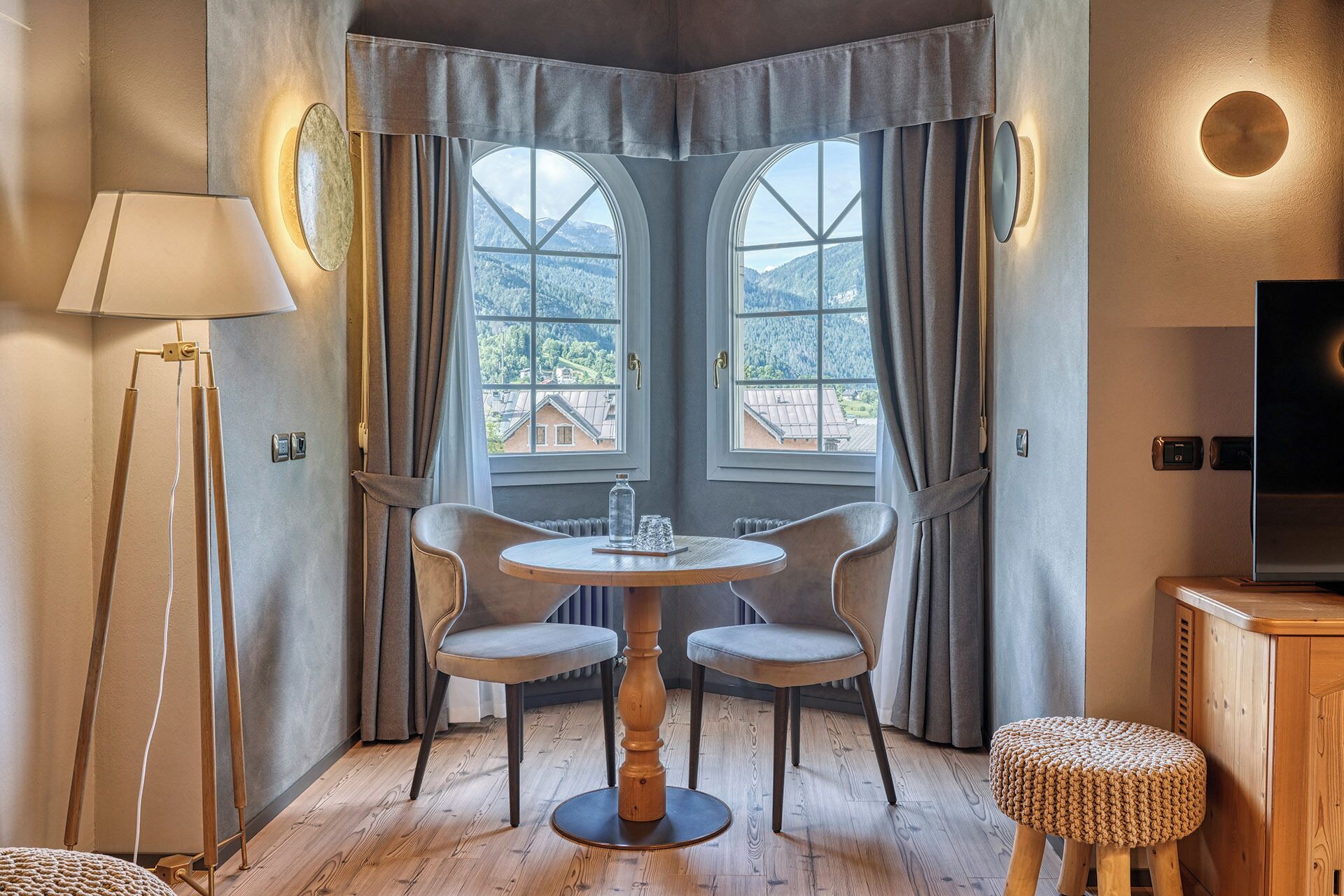 A corner room with two large windows, a round wooden table with two chairs, a floor lamp, a small stool, and a television on a cabinet.