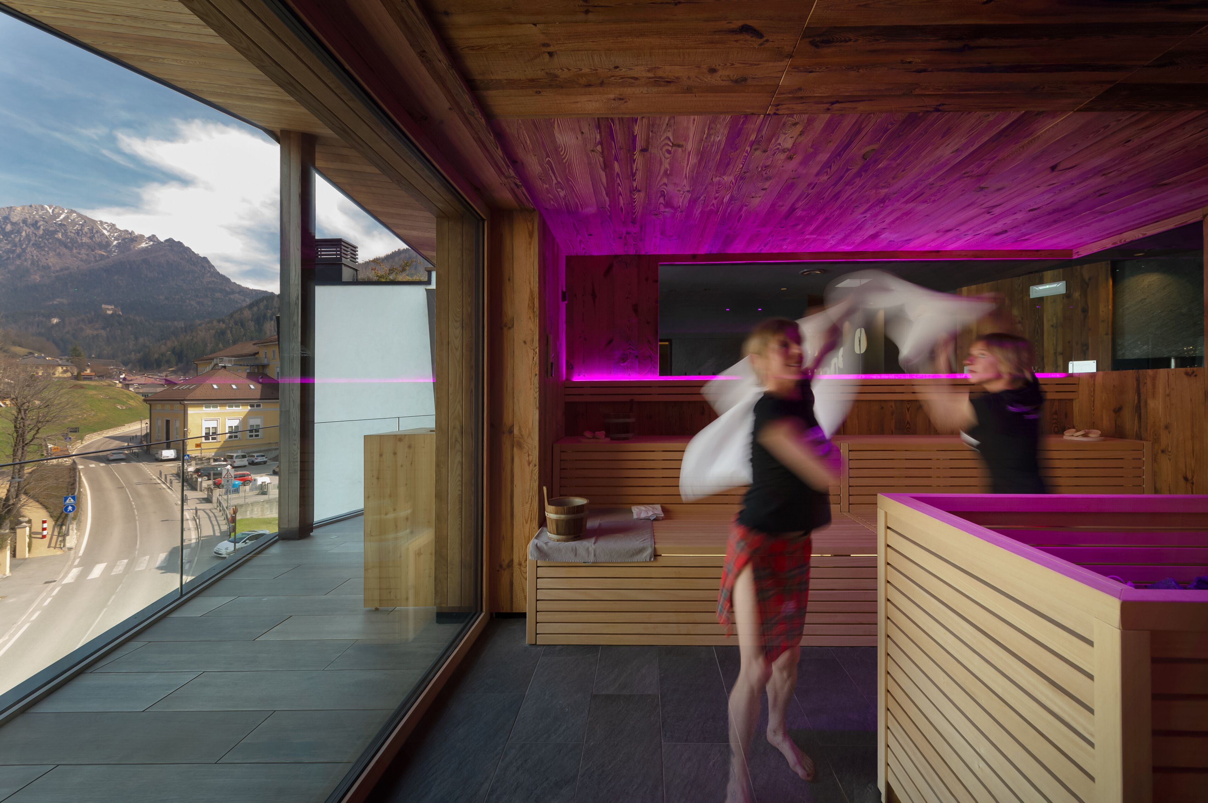 A modern spa room with wooden flooring, a large window overlooking a road and mountains, and a wooden bench with a small table.
