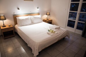A neatly made bed with white linens, two bedside lamps, a wooden nightstand, and a window with a view of the outside. - Lefkothea (Santorini)