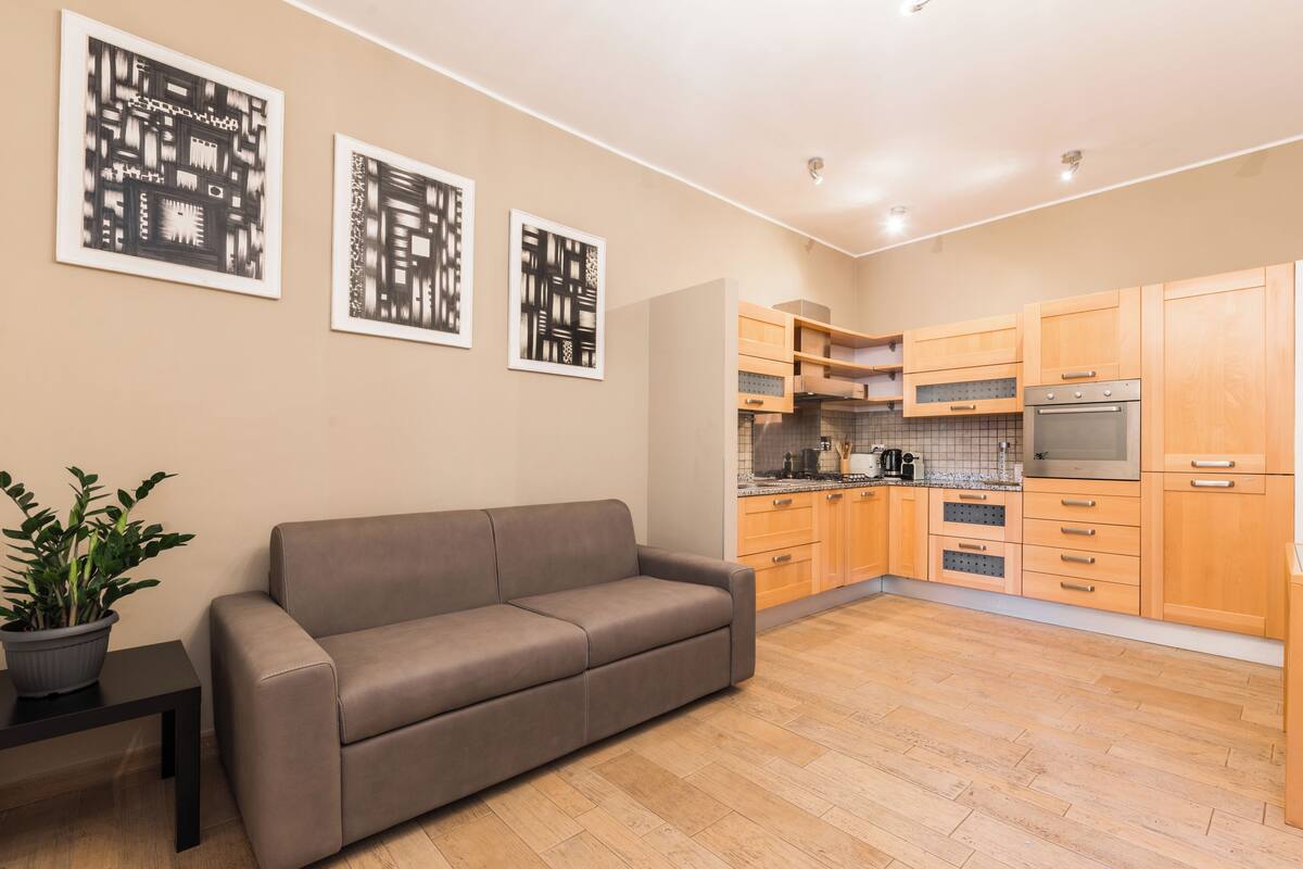 Ein modernes Wohnzimmer mit einer Couch, einem kleinen Tisch mit Topfpflanze und einer Küche mit Holdekor und Geräten.