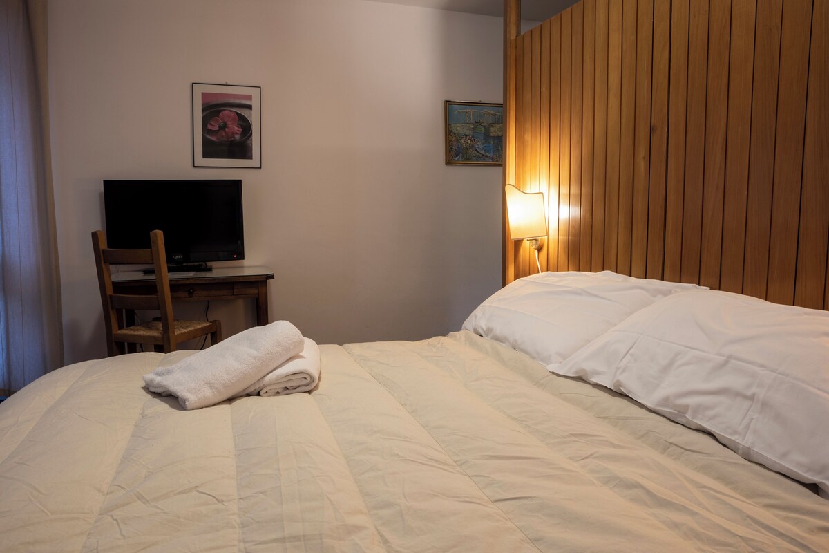 A neatly made bed with a folded towel, a television on a wooden desk, and two wall-mounted lamps.