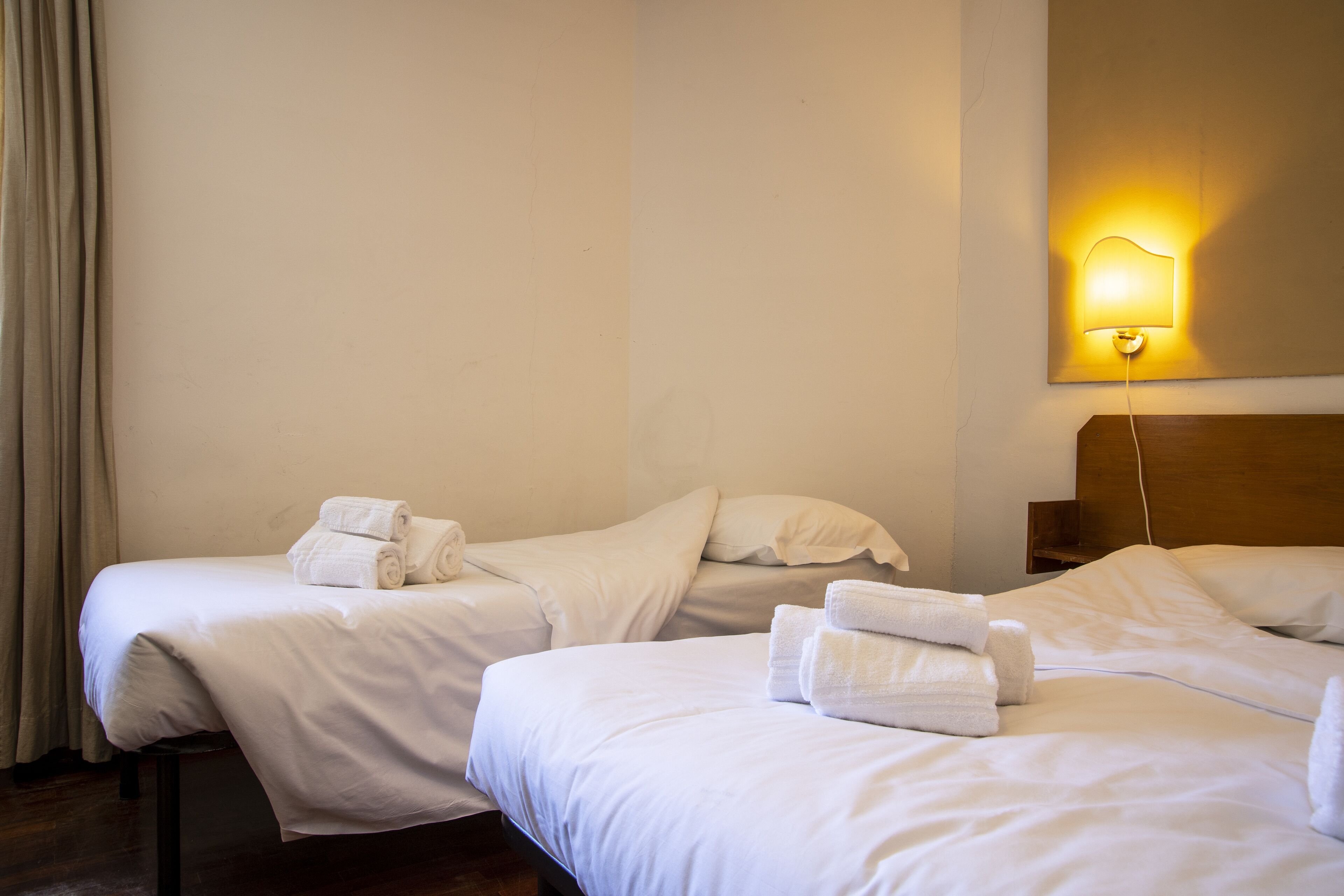 Two single beds with white linens and towels, a wooden headboard, and a wall-mounted light fixture.
