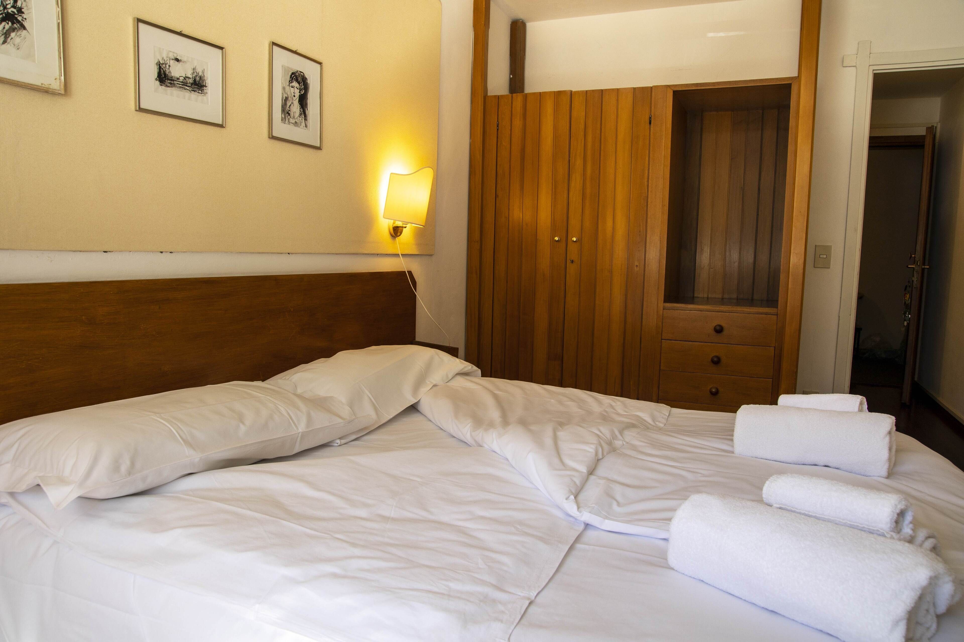 A neatly made bed with white linens, a wooden wardrobe, and a wall-mounted light fixture.