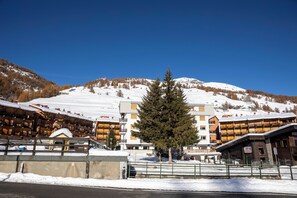 Un hotel de varios pisos, con una pista de esquí al fondo y un árbol destacado en primer plano.