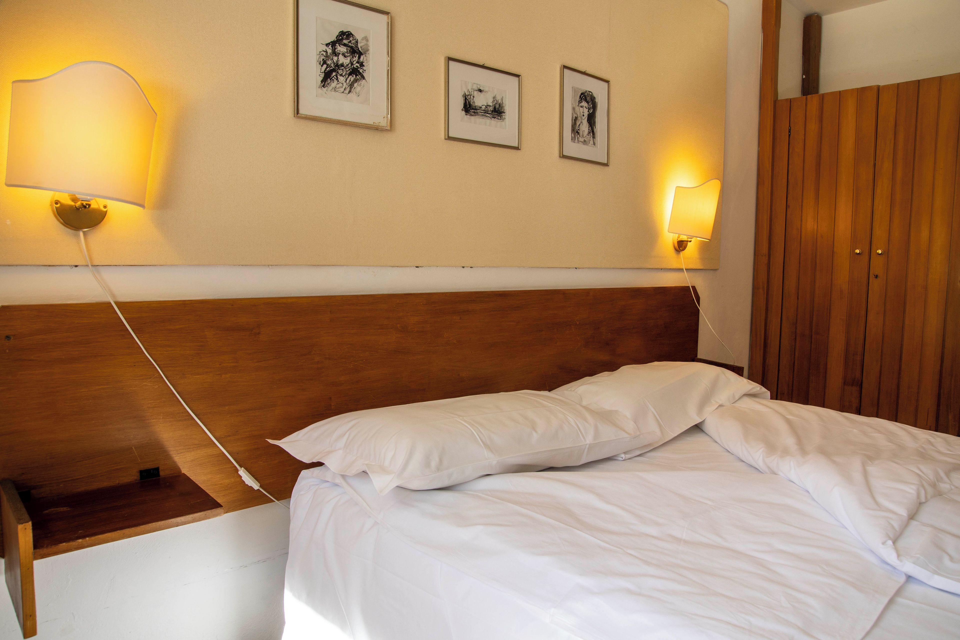 Een netjes opgemaakt bed met witte lakens, een houten hoofdbord en een nachtkastje met een lamp. Boven het bed hangen drie ingelijste foto's aan de muur.