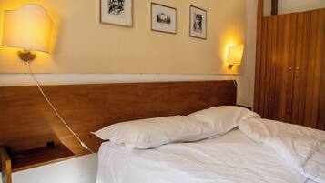 Een netjes opgemaakt bed met witte lakens, een houten hoofdbord en een nachtkastje met een lamp. Boven het bed hangen drie ingelijste foto's aan de muur.
