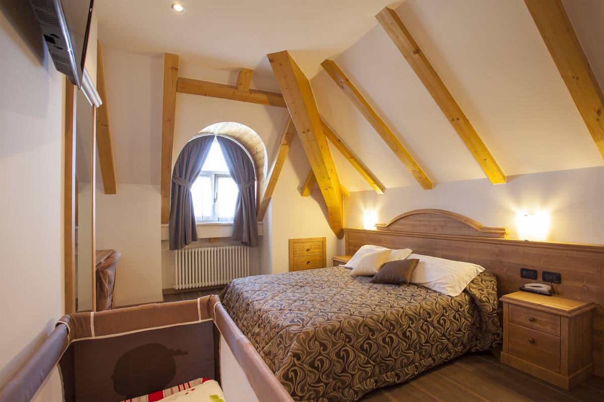 A bedroom with a sloped ceiling, wooden beams, a bed with patterned bedding, a nightstand, and a bedside table with a lamp.