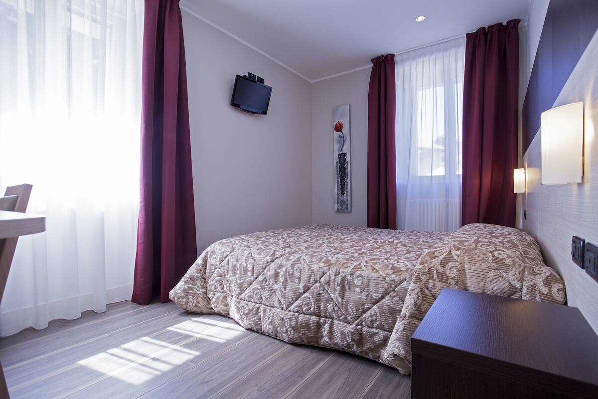 A bedroom with a bed, a bedside table, a wall-mounted TV, and a window with curtains.
