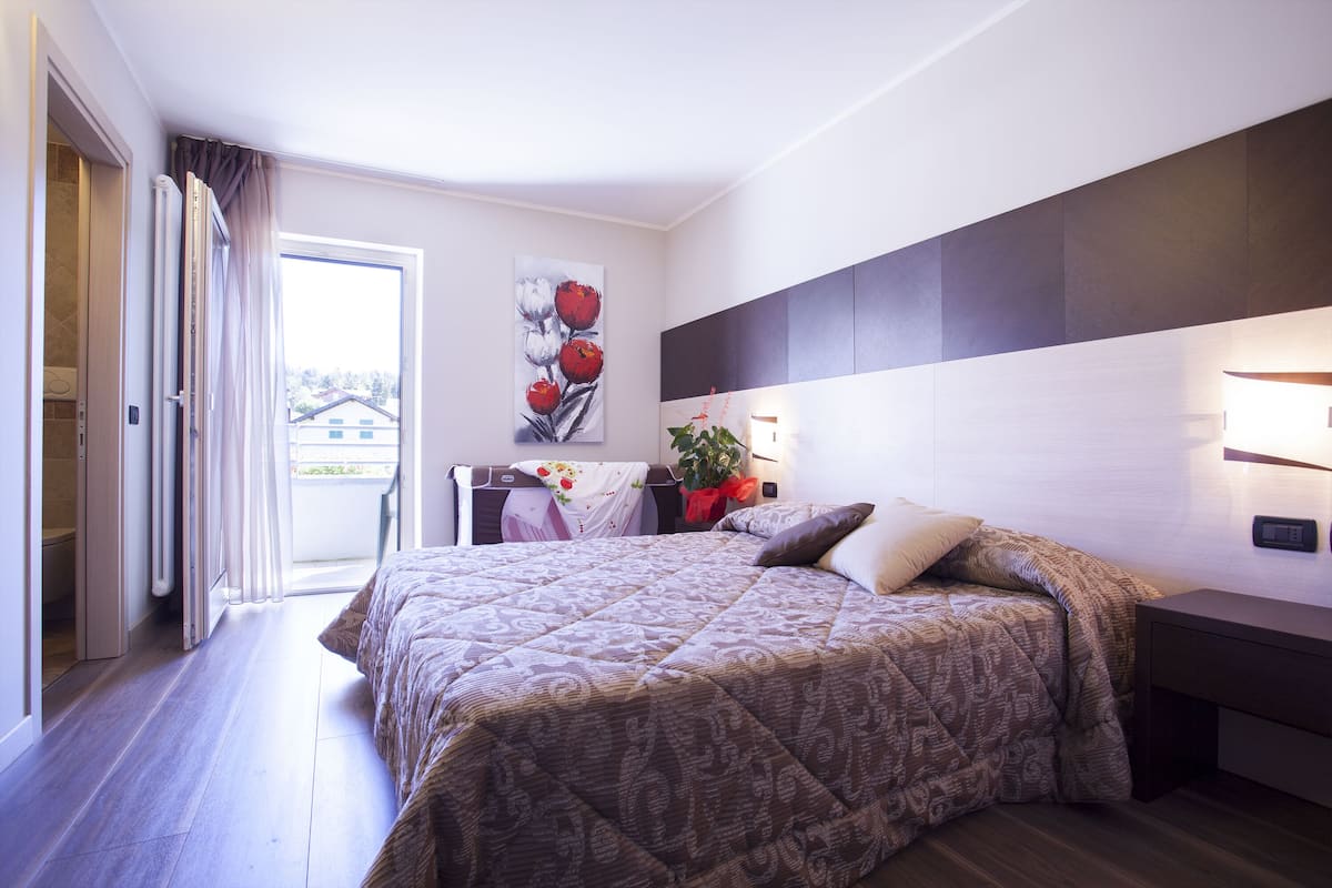 A hotel room with a large bed, a bedside table, a nightstand, a wall-mounted lamp, a painting, and a view through the window.